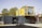 A structure made from shipping containers is set up outdoors. The lower container is dark gray and functions as the main space, featuring a door and large windows. On top sits a yellow container with large, clear windows, likely serving as an additional room or office space. A picnic table and a bicycle are located in front of the structure. Tall trees and a brick building are visible in the background under a clear sky.