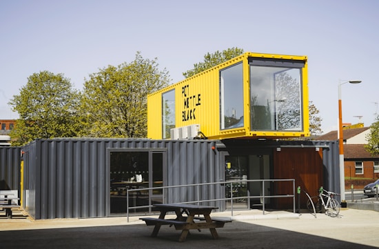 A structure made from shipping containers is set up outdoors. The lower container is dark gray and functions as the main space, featuring a door and large windows. On top sits a yellow container with large, clear windows, likely serving as an additional room or office space. A picnic table and a bicycle are located in front of the structure. Tall trees and a brick building are visible in the background under a clear sky.