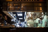 A professional kitchen setting featuring hanging kitchen utensils against a green tiled wall. Shelves stocked with bottles and jars are visible above, while a person is seen in the background, presumably working. A vase with flowers is placed on a counter, creating a contrast between the industrial environment and a touch of nature.