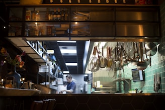 A professional kitchen setting featuring hanging kitchen utensils against a green tiled wall. Shelves stocked with bottles and jars are visible above, while a person is seen in the background, presumably working. A vase with flowers is placed on a counter, creating a contrast between the industrial environment and a touch of nature.