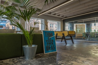 A modern office space with a large green plant in a metallic pot, a motivational sign saying 'Think of Your Own Ideas', and a foosball table. The area has large windows and a glass conference room visible in the background. The interior includes a mix of natural and industrial elements, with visible ceiling beams and a polished concrete floor.