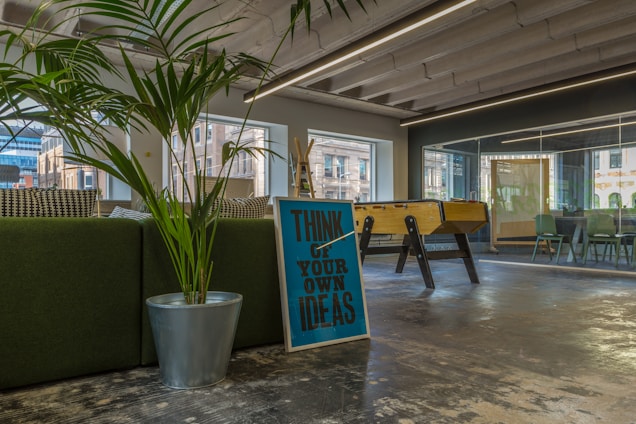 A modern office space with a large green plant in a metallic pot, a motivational sign saying 'Think of Your Own Ideas', and a foosball table. The area has large windows and a glass conference room visible in the background. The interior includes a mix of natural and industrial elements, with visible ceiling beams and a polished concrete floor.