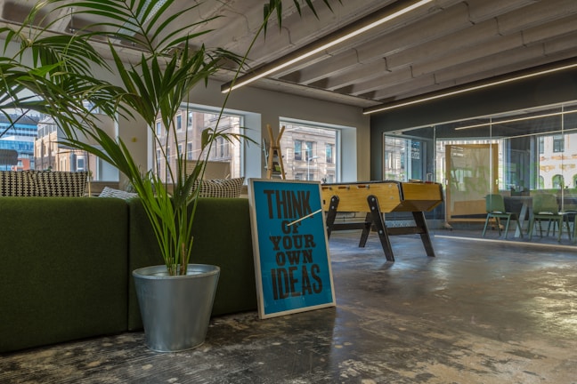 A modern office space with a large green plant in a metallic pot, a motivational sign saying 'Think of Your Own Ideas', and a foosball table. The area has large windows and a glass conference room visible in the background. The interior includes a mix of natural and industrial elements, with visible ceiling beams and a polished concrete floor.