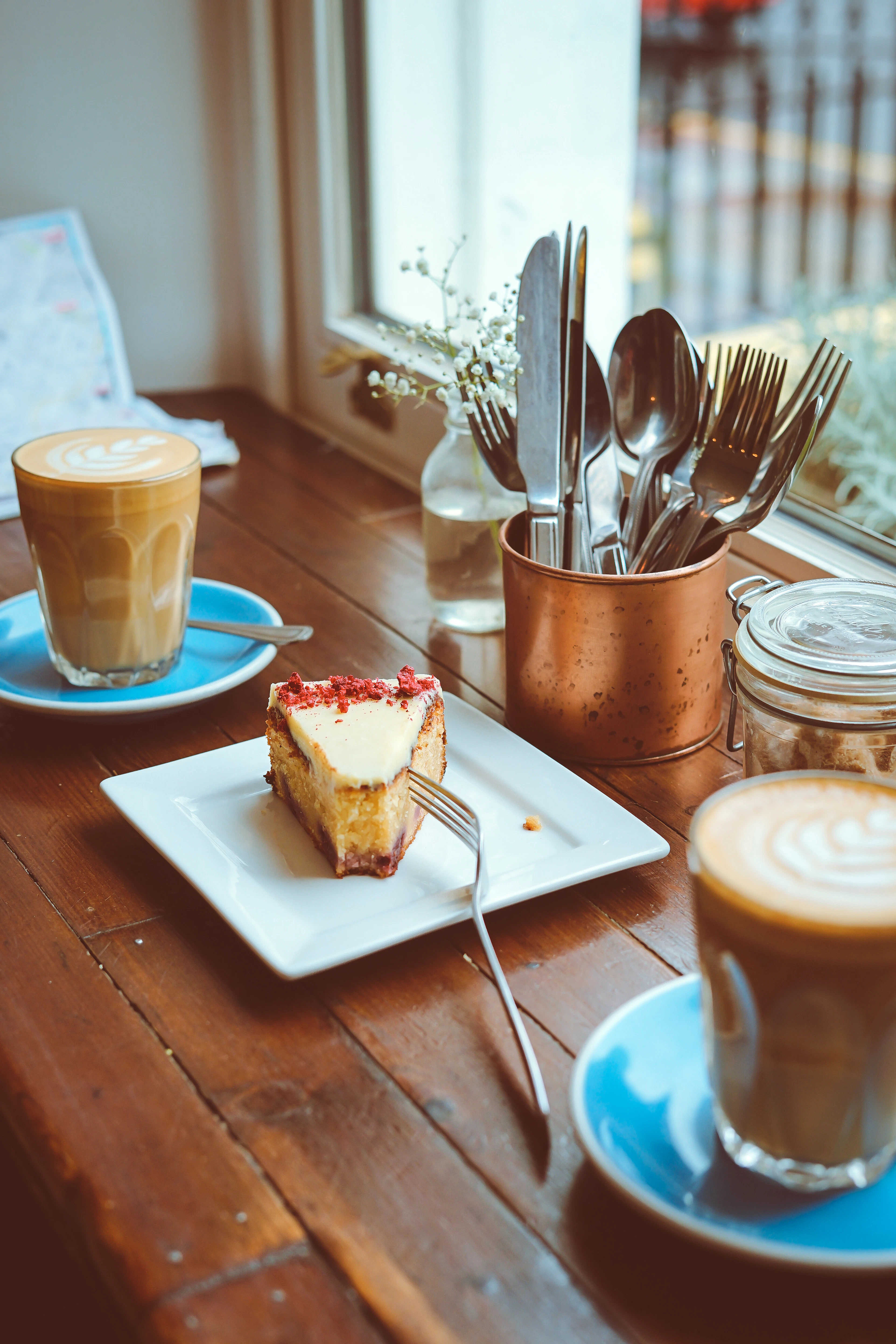 A latte and a slice of cheesecake on a cafe table with cutlery in a copper pot.