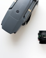 A grey, foldable drone with visible propellers rests on a white surface next to a black camera lens with intricate markings and focus numbers.