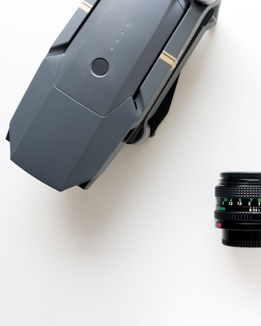 A grey, foldable drone with visible propellers rests on a white surface next to a black camera lens with intricate markings and focus numbers.