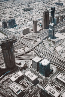 An aerial view of a sprawling urban landscape dominated by a complex network of intersecting highways and overpasses. Tall skyscrapers and various commercial buildings are situated throughout the cityscape, creating a dense and modern urban environment. Surrounding the central infrastructure are smaller residential areas and patches of greenery.