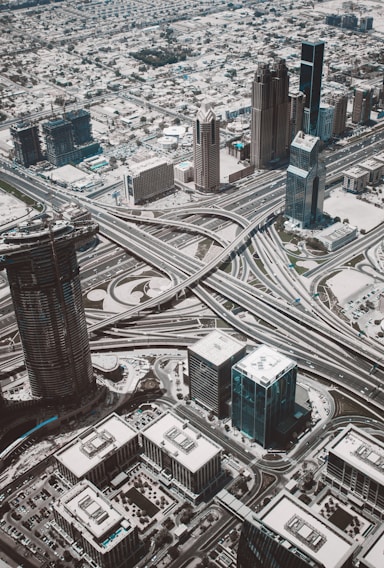An aerial view of a sprawling urban landscape dominated by a complex network of intersecting highways and overpasses. Tall skyscrapers and various commercial buildings are situated throughout the cityscape, creating a dense and modern urban environment. Surrounding the central infrastructure are smaller residential areas and patches of greenery.