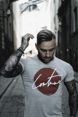 A man with tattoos on his arms, wearing a t-shirt featuring a large red circle with the word 'couture' written across it. He stands in a narrow alleyway with stone pavement, framed by tall buildings on either side. His hand is raising to adjust his hair.