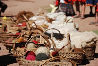 A row of woven baskets filled with various colorful yarns and wool is arranged on the ground in an outdoor setting. In the background, there are people standing, partially visible from the waist down. The baskets hold yarns of different colors including reds, greens, and beiges, and there are tools for wool spinning visible among the materials.