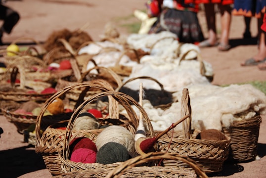 A row of woven baskets filled with various colorful yarns and wool is arranged on the ground in an outdoor setting. In the background, there are people standing, partially visible from the waist down. The baskets hold yarns of different colors including reds, greens, and beiges, and there are tools for wool spinning visible among the materials.