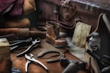 A collection of vintage cobbler tools and materials is arranged on a workbench. Tools like pliers and a hammer are scattered among leather pieces and a small, finely crafted boot. An old suitcase and a faded drawing are also present, suggesting a traditional shoe repair setup.