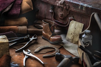 A collection of vintage cobbler tools and materials is arranged on a workbench. Tools like pliers and a hammer are scattered among leather pieces and a small, finely crafted boot. An old suitcase and a faded drawing are also present, suggesting a traditional shoe repair setup.