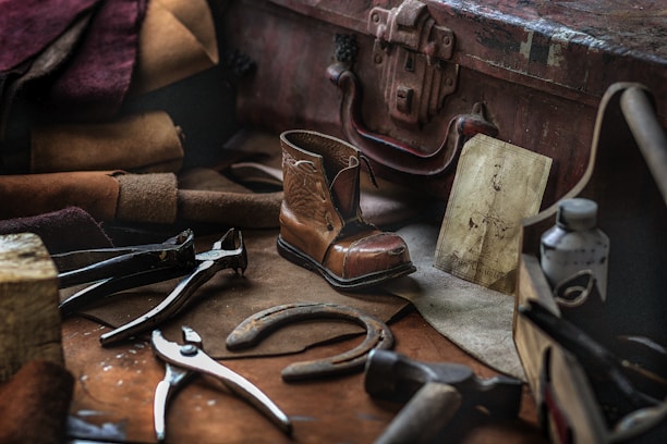 A collection of vintage cobbler tools and materials is arranged on a workbench. Tools like pliers and a hammer are scattered among leather pieces and a small, finely crafted boot. An old suitcase and a faded drawing are also present, suggesting a traditional shoe repair setup.