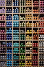 A neatly organized display of colorful spray paint cans is arranged in a grid pattern within individual cubbies. The cans are grouped by color, featuring a wide spectrum including blue, green, yellow, orange, red, and purple. Each compartment holds a tightly packed collection of spray cans, creating a vibrant and orderly visual effect.