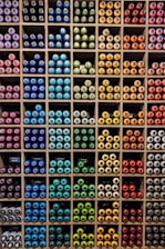 A neatly organized display of colorful spray paint cans is arranged in a grid pattern within individual cubbies. The cans are grouped by color, featuring a wide spectrum including blue, green, yellow, orange, red, and purple. Each compartment holds a tightly packed collection of spray cans, creating a vibrant and orderly visual effect.