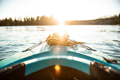 A kayak's bow points towards a bright, sunlit horizon over a calm body of water, with coiled rope resting on top. Trees line the distant shoreline under a clear sky.
