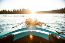 A kayak's bow points towards a bright, sunlit horizon over a calm body of water, with coiled rope resting on top. Trees line the distant shoreline under a clear sky.