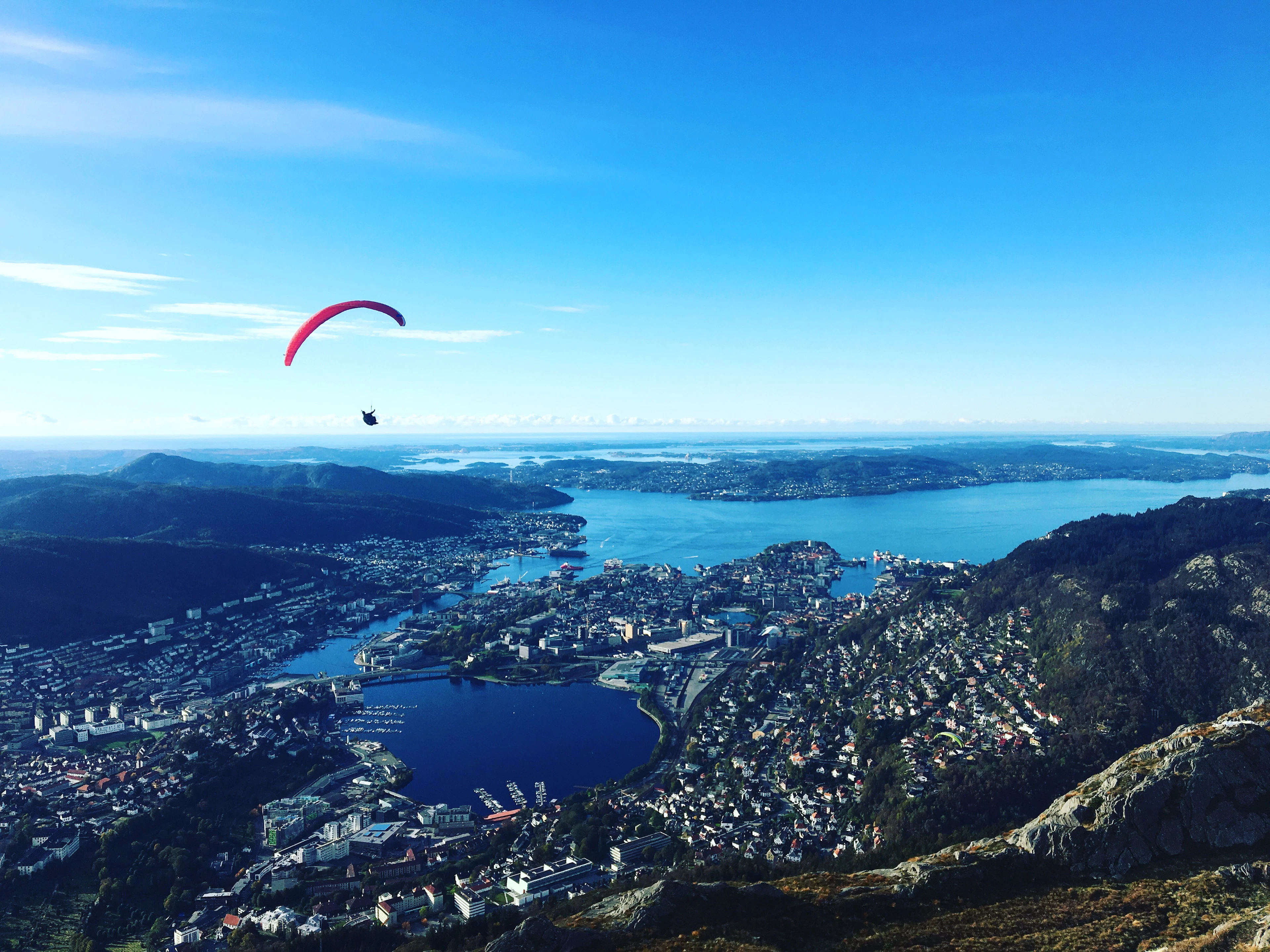 A pink paraglider hanging in a deep blue sky above Bergen, seen from the summit of Ulriken, with the city, its dark harbour lake and the fjord threading between wooded islands spread out far below.