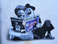 Graffiti art on a wall depicting two figures with long beards reading a copy of Charlie Hebdo. The figures are stylized and humorously portrayed, sitting comfortably on the ground with the newspaper prominently displayed. The artwork exhibits a blend of black, white, and shades of gray with touches of purple and blue accents.