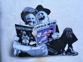 Graffiti art on a wall depicting two figures with long beards reading a copy of Charlie Hebdo. The figures are stylized and humorously portrayed, sitting comfortably on the ground with the newspaper prominently displayed. The artwork exhibits a blend of black, white, and shades of gray with touches of purple and blue accents.