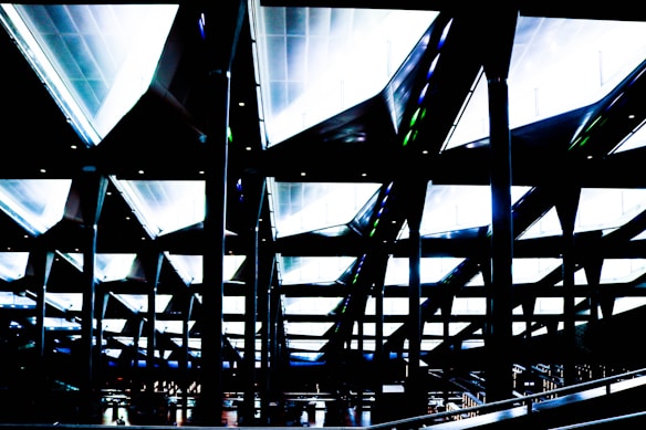 Futuristic architectural ceiling design featuring a series of intersecting lines and angled beams illuminated by bright lights. The composition creates a geometric and almost surreal visual effect.