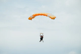 A vibrant shot of a skydiver freefalling with a bright orange parachute against a clear blue sky.