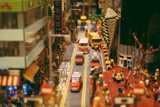 A bustling street scene made with LEGO bricks, featuring multiple vehicles on a road lined with buildings. LEGO figures are gathered along the sidewalks, resembling a lively urban environment.