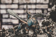 A detailed miniature scene depicts a warrior in ornate armor riding a dragon-like creature with intricate scales and a long tail. The warrior is raising a sword triumphantly. Other smaller figures, also clad in armor, surround the main figure, appearing as if ready for battle. The backdrop is blurred, emphasizing the detailed craftsmanship of the models.