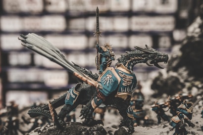 A detailed miniature scene depicts a warrior in ornate armor riding a dragon-like creature with intricate scales and a long tail. The warrior is raising a sword triumphantly. Other smaller figures, also clad in armor, surround the main figure, appearing as if ready for battle. The backdrop is blurred, emphasizing the detailed craftsmanship of the models.