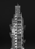 A tall, multi-level construction structure covered in scaffolding. The building appears incomplete, with various platforms and support beams visible. The contrasting light and dark elements highlight the intricate framework against a solid dark background.