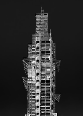 A tall, multi-level construction structure covered in scaffolding. The building appears incomplete, with various platforms and support beams visible. The contrasting light and dark elements highlight the intricate framework against a solid dark background.
