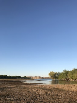 A serene landscape featuring a wide, open sky with a bright blue hue. A calm body of water stretches across the middle of the image, bordered by patches of sandy, dry land. On the right side, clusters of green trees add a touch of nature, and in the distance, low hills provide a subtle backdrop.