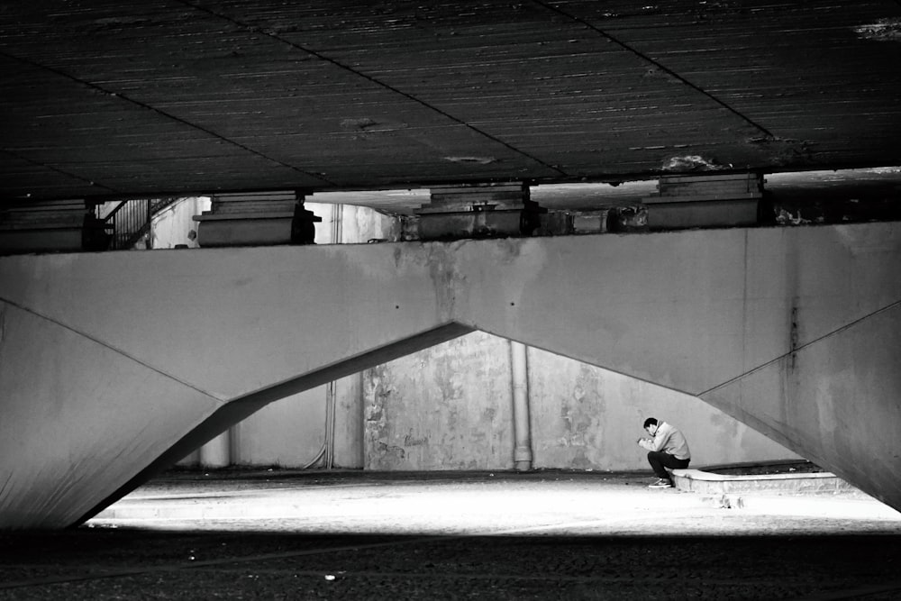 grayscale photography of man sitting under the bridge