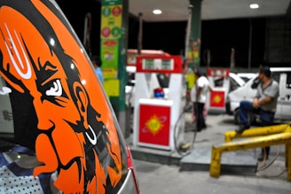 A gas station at night with multiple fuel pumps and cars being refueled. On the left side, a bold orange graphic of a face is prominently displayed on a vehicle. Two people are present; one appears to be attending to a pump while another is sitting on a railing.