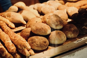 A variety of freshly baked bread and pastries are displayed on a wooden shelf. There are different types of bread rolls, long breadsticks, and some round loaves with a rustic appearance. The baked goods have a golden-brown crust, indicating a well-baked texture.