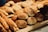 A variety of freshly baked bread and pastries are displayed on a wooden shelf. There are different types of bread rolls, long breadsticks, and some round loaves with a rustic appearance. The baked goods have a golden-brown crust, indicating a well-baked texture.