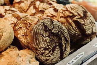 Various loaves of bread with rough crust patterns, displayed on a counter in a bakery setting. The bread appears freshly baked with a rustic appearance, and a handwritten label indicates they might be rye walnut loaves.