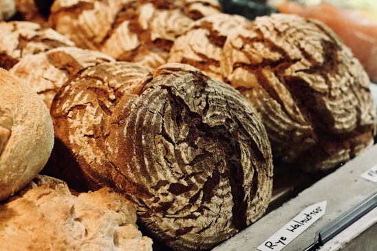 Various loaves of bread with rough crust patterns, displayed on a counter in a bakery setting. The bread appears freshly baked with a rustic appearance, and a handwritten label indicates they might be rye walnut loaves.