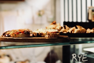 A rustic wooden board holds slices of bread topped with various ingredients, including tomatoes and a white spread. The background features a display of more baked goods, possibly croissants or pastries. The word 'RYE' is visible on the glass surface below the board.