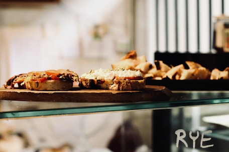 A rustic wooden board holds slices of bread topped with various ingredients, including tomatoes and a white spread. The background features a display of more baked goods, possibly croissants or pastries. The word 'RYE' is visible on the glass surface below the board.