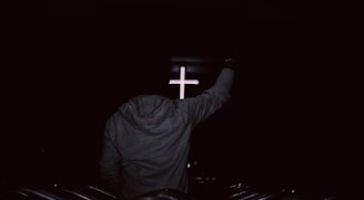 A person wearing a hoodie is standing with an arm raised in front of a softly illuminated cross, set against a dark background, suggesting a spiritual or religious environment.