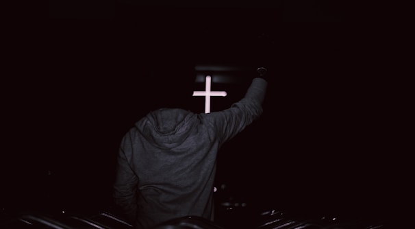 A person wearing a hoodie is standing with an arm raised in front of a softly illuminated cross, set against a dark background, suggesting a spiritual or religious environment.
