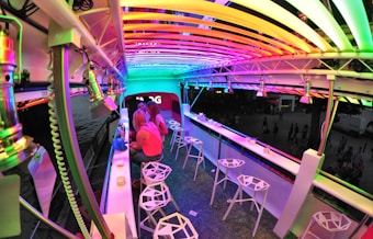 A bar setting with modern, white geometric stools and a sleek counter. The area is illuminated by vibrant, multicolored LED lights creating a lively and futuristic atmosphere.