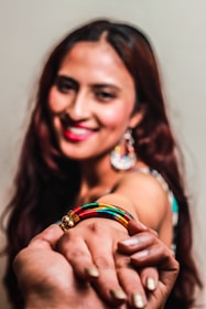 A smiling woman with long, dark hair holds out her hand adorned with a colorful bracelet featuring yellow, green, red, and black segments. She wears a pair of colorful, dangling earrings and has red lipstick. Another person's hand is gently holding her hand.