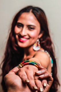 A smiling woman with long, dark hair holds out her hand adorned with a colorful bracelet featuring yellow, green, red, and black segments. She wears a pair of colorful, dangling earrings and has red lipstick. Another person's hand is gently holding her hand.