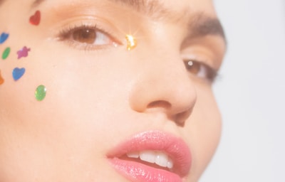 A close-up of a person's face adorned with colorful adhesive shapes, such as hearts and stars, near the eye and cheek. The skin is smooth and the lighting is soft, creating a gentle highlight on the face. The lips are slightly parted and have a glossy finish, adding to the overall softness and vibrancy of the image.