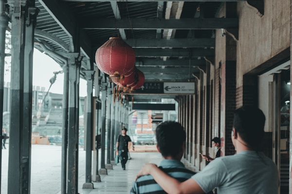 A covered walkway with red lanterns hanging from the ceiling. Two people in the foreground are walking with one person’s hand on the other's shoulder, creating a friendly gesture. In the background, a person is sitting and looking at a phone or small device. The setting appears to be in an urban area with elements of traditional and modern architecture.