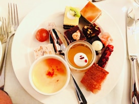 An assortment of desserts on a white plate, including various small cakes, a bowl of crème brûlée, and a small glass of a yellow dessert topped with a white dollop. The plate is accompanied by a spoon and cutlery on a white tablecloth.
