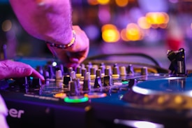 A person is adjusting controls on a DJ mixer and turntable, suggesting they are mixing music. The scene is illuminated with colorful lights, creating a vibrant and energetic atmosphere.